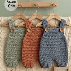 May include: Three handmade crochet baby rompers in sage green, burnt orange, and dusty blue, each with wooden button closures. The rompers hang on wooden hangers with the text "Made with Love". A sign reads "Pattern Only".