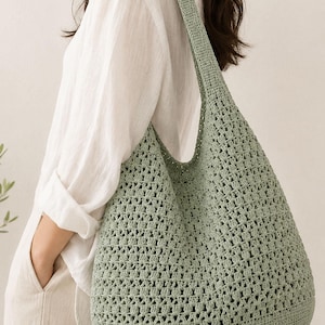 Può includere: Una borsa hobo all'uncinetto color verde salvia con un motivo ripetuto a piastrelle granny e un'ampia tracolla. La borsa presenta una base solida e un testo circolare sovrapposto nell'angolo in alto a sinistra con la scritta Pattern Only.