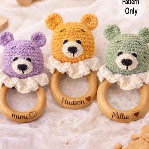 May include: Three handmade crochet bear baby rattles in purple, yellow, and green. Each bear has a black nose and is attached to a wooden ring with a name engraved on it. The rattles are on a white, textured surface.