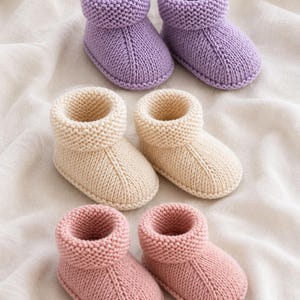 May include: Three pairs of hand-knitted baby booties in soft pastel colors. The top pair is lavender, the middle pair is cream, and the bottom pair is dusty pink. Each pair features a garter stitch cuff and a smooth stockinette stitch foot.