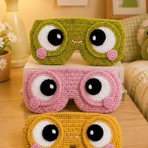 Puede incluir: Tres antifaces de crochet en verde, rosa y amarillo. Cada antifaz presenta ojos grandes blancos y negros, una boca pequeña bordada y dos mejillas circulares rosas. Los antifaces están apilados verticalmente, mostrando un diseño uniforme.