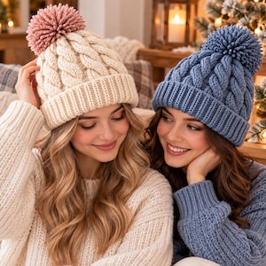 Puede incluir: Dos gorros de punto con pompones. Uno es de color crema con un pompón rosa empolvado y el otro es azul con un pompón azul a juego. Ambos gorros presentan un grueso patrón de punto de ochos y un ancho borde acanalado.