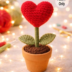 Puede incluir: Una flor en forma de corazón tejida a ganchillo en una maceta de terracota. El corazón rojo florece sobre un tallo verde con dos hojas verdes. La maceta está llena de hilo marrón, y la imagen incluye el texto "Pattern Only".