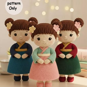 Op de afbeelding: Drie handgemaakte gehaakte poppen met bruine knotjes versierd met bloemen. Elke pop draagt een andere kleur jurk: blauwgroen, roze en groen. De poppen hebben geborduurde gezichtskenmerken en staan naast elkaar. De tekst "pattern Only" staat in een witte cirkel.