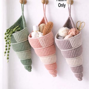 May include: Three handmade crochet shell-shaped hanging baskets in shades of green, pink, and purple. The baskets are suspended from wooden pegs on a white wall. One basket contains a trailing plant, another toiletries, and the third, yarn and scissors. Text reads "Pattern Only".