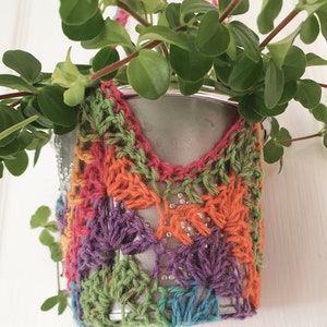 Puede incluir: Una pequeña planta en una maceta de metal con una cubierta de ganchillo de colores. La cubierta está hecha con hilo de diferentes colores en un patrón de arcoíris.
