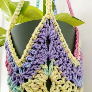May include: A crocheted plant hanger with a rainbow colour scheme. The hanger is made of yarn in shades of pink, purple, blue, green, and yellow. The hanger is designed to hold a small plant pot.