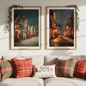 以下が含まれることがあります： 雪が降る街並みの絵が2枚、クリーム色のソファの上に飾られています。赤いチェック柄のクッションと「JOY」の刺繍が入ったクッションがソファを飾り、居心地の良い休日の雰囲気を演出しています。
