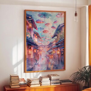 以下が含まれることがあります： 額縁に入った水彩画は、建物、提灯、カラフルな傘が空に吊り下げられた日本の街並みを描いています。 このアートワークは、ピンク、青、オレンジの柔らかな色使いで、穏やかな雰囲気を作り出しています。 絵は白い壁に飾られています。
