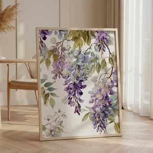 以下が含まれることがあります： 紫、ラベンダー、緑の色合いの藤の花が描かれた、額入りの水彩画プリント。アートワークは白い背景にあり、明るい木製のフレームに入っています。プリントは、自然光が入る部屋の壁に立てかけられています。