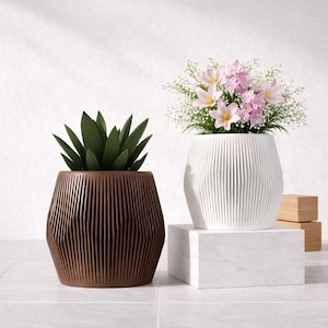 Peut inclure: Deux pots décoratifs avec un design côtelé. L'un est marron et contient une plante succulente verte, l'autre est blanc et affiche des fleurs roses. Le pot blanc repose sur un bloc de marbre blanc. Idéal pour la décoration intérieure.