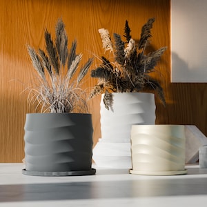 May include: Three decorative ceramic planters in shades of gray, white, and cream, each with a wavy, textured design. The planters are filled with dried ornamental grasses, creating a natural and stylish display. The planters are sitting on a white surface.