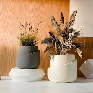 May include: Two cylindrical planters with a diagonal wrap-around design. One planter is matte charcoal gray and holds thin, upright green grasses. The other is matte cream and contains a cluster of feathery, dried beige and brown botanical stems.