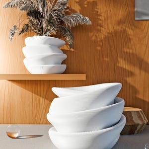 Peut inclure: Une pile de quatre bols blancs de forme organique avec une surface texturée. Un vase blanc assorti contient des herbes ornementales séchées. L'arrière-plan présente un mur en bois et une petite table d'appoint en bois.