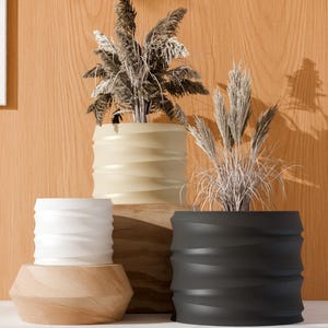 May include: Four decorative planters in different colours and styles. The planters are beige, white, black, and natural wood. Each planter has a unique design, with wavy or geometric patterns. The planters are filled with dried ornamental grasses, creating a natural and modern aesthetic.