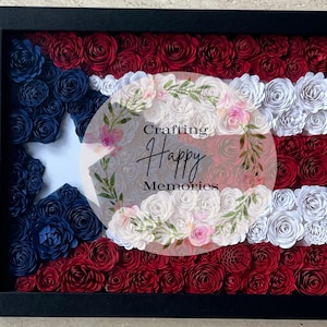 May include: A framed artwork featuring a floral arrangement in the style of the Puerto Rican flag. The design incorporates red, white, and blue paper flowers, with a white star and the text "Crafting Happy Memories". The frame is black.