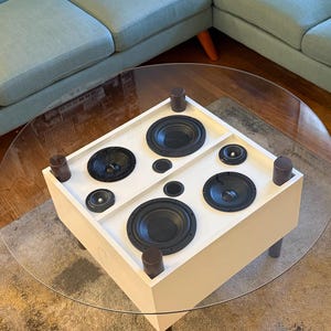Op de afbeelding: Een witte, vierkante salontafel met een doorzichtig glazen blad. De tafel heeft vier grote zwarte luidsprekers en twee kleinere. De tafel wordt omringd door een lichtblauwe hoekbank met gele kussens en een patroonkleed.