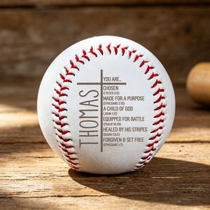 May include: A white baseball with red stitching and the name "THOMAS" printed vertically. The baseball has a cross and inspirational text including "CHOSEN" and "MADE FOR A PURPOSE". A wooden baseball bat is in the background.