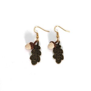May include: A pair of crochet earrings with a leaf and acorn design. The earrings have gold-coloured ear wires. The leaves are dark green, and the acorns are beige and brown. The earrings are set against a white background.