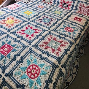 Puede incluir: Una colcha hecha a mano, tejida a ganchillo, con un patrón repetitivo de diseños de estrellas coloridas. La colcha muestra una paleta de blanco, azul, rosa, amarillo y verde, con cada cuadrado enmarcado por un borde azul oscuro. La colcha está sobre una cama.