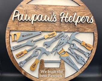 Op de afbeelding: Een rond houten bord met de tekst "Pawpaw's Helpers" en "We built the best Pawpaw!". Het heeft houten uitsparingen van gereedschappen, elk met een naam zoals Maddox, Sage en Sophia. Het bord heeft een lichtgekleurd middengedeelte.