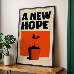 Può includere: Stampa artistica incorniciata con le parole "A NEW HOPE" in grassetto nero. Sotto, un rettangolo arancione con l'illustrazione di una pianta in vaso. La stampa è in una cornice nera e poggia su una superficie di legno.