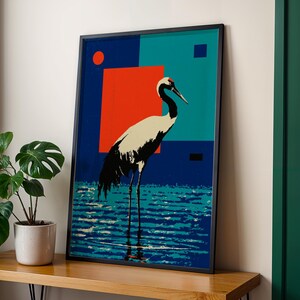 Op de afbeelding: Ingelijste kunstdruk met een gestileerde kraanvogel in water. Het kunstwerk heeft een geometrische achtergrond met rode, turquoise en marineblauwe blokken. De kraanvogel is afgebeeld in zwart en wit met een rode kop. De print zit in een zwart frame.
