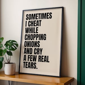 Könnte beinhalten: Ein gerahmter Kunstdruck mit dem Text "SOMETIMES I CHEAT WHILE CHOPPING ONIONS AND CRY A FEW REAL TEARS" in schwarzer Schrift. Der Druck befindet sich in einem schwarzen Rahmen und steht auf einer Holzoberfläche. Links ist eine Topfpflanze.