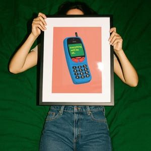 Könnte beinhalten: Ein gerahmter Druck mit einer Illustration eines blauen Mobiltelefons auf korallenfarbenem Hintergrund. Auf dem Display steht der grüne Text "everything will be ok." Das Gerät hat eine schwarze Antenne, rote Tasten und ein Tastenfeld.