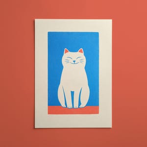 Könnte beinhalten: Ein rechteckiger Kunstdruck mit einer stilisierten weißen Katze mit lächelndem Gesichtsausdruck vor einem einfarbigen blauen Hintergrund. Die Katze sitzt auf einem dünnen roten horizontalen Streifen und ist von einem breiten cremefarbenen Rand umgeben.