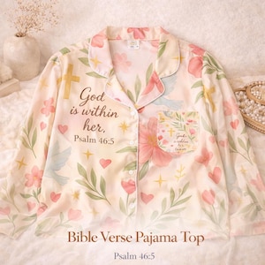 Puede incluir: Una parte superior de pijama de color claro con un estampado floral y de cruces. La parte superior presenta el texto "God is within her, Psalm 46:5" y "Bible Verse Pajama Top, Psalm 46:5". El diseño incluye flores rosas, corazones y palomas.