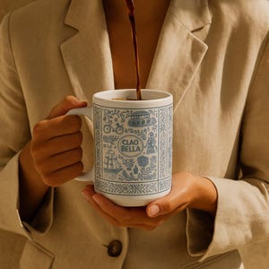 Puede incluir: Taza de cerámica blanca con un diseño decorativo azul y blanco y el texto "CIAO BELLA". La taza se está llenando con un líquido oscuro. El diseño incluye imágenes de una bicicleta, un barco y un edificio.