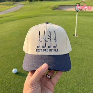 May include: A beige and navy blue baseball cap with the text "BEST DAD BY PAR" and golf club graphics. The cap has the names "James", "John", "Rachel", and "William" embroidered on it. The cap is being held on a golf course.