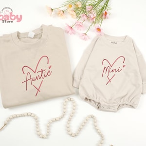 May include: Beige Auntie and Mini matching set. The adult sweatshirt and baby romper feature a red heart design with the words "Auntie" and "Mini" in a script font. The set is displayed on a white surface with floral accents and a beaded garland.