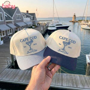 May include: Two baseball caps with the text "CAPE COD" and a cocktail graphic. The caps have a cream-colored crown and a gray and navy blue brim. The text "Est. 2026 Social Club" is also printed on the caps. The background shows a harbor scene.
