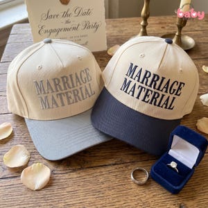 May include: Two baseball caps with "MARRIAGE MATERIAL" embroidered on the front. One cap is tan with a gray brim, the other is tan with a navy brim. A ring box with a diamond ring and a wedding band are also visible.