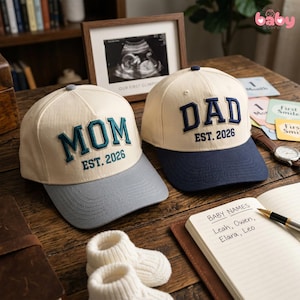 May include: Two baseball caps, one reading "MOM EST. 2026" with a gray brim, and the other "DAD EST. 2026" with a navy brim. The caps sit on a wooden surface with baby booties, a framed sonogram, and a notebook with baby names.