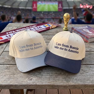 May include: Two baseball caps with cream-colored crowns and embroidered text reading "I am from Bosnia take me to America." One cap features a light gray brim, while the other has a dark navy blue brim. Both caps have a structured five-panel design.