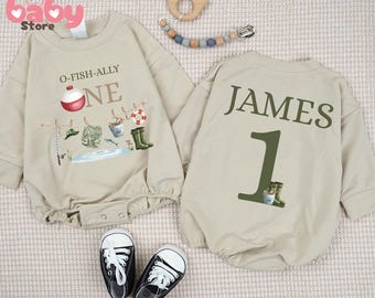 May include: Beige baby bodysuit with long sleeves. The front features the text "O-FISH-ALLY ONE" with fishing-themed graphics. The back has the name "JAMES" and the number "1". Black and white baby sneakers are at the bottom.