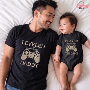 May include: Matching black t-shirts and onesie with video game controller graphics. The adult shirt says "LEVELED up to DADDY." The baby's onesie says "PLAYER has entered THE GAME."