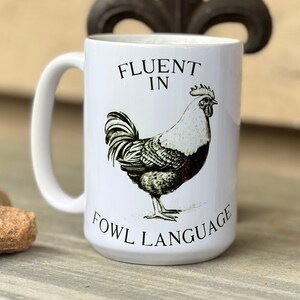 May include: White ceramic coffee mug with a black and white illustration of a rooster and the text "Fluent in Fowl Language".
