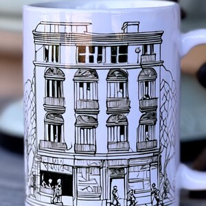 May include: White ceramic mug featuring a black and white illustration of a building with multiple windows and a shop front. The building is drawn in a simple, stylised manner. There are small figures of people walking in front of the building.