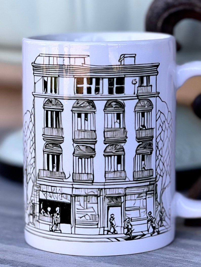May include: White ceramic mug featuring a black and white illustration of a building with multiple windows and a shop front. The building is drawn in a simple, stylised manner. There are small figures of people walking in front of the building.