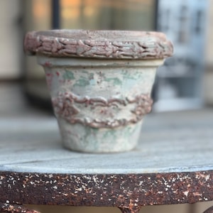 May include: A weathered terracotta plant pot featuring a distressed finish with patches of faded green and white paint. The exterior is decorated with a raised, molded relief pattern around the rim and a central band of ornate, scrolling floral motifs.