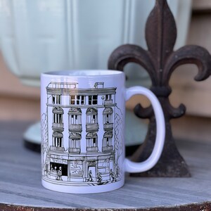 May include: White ceramic mug featuring a black and white illustration of a building with windows and balconies. The building is surrounded by trees and people walking on the pavement.
