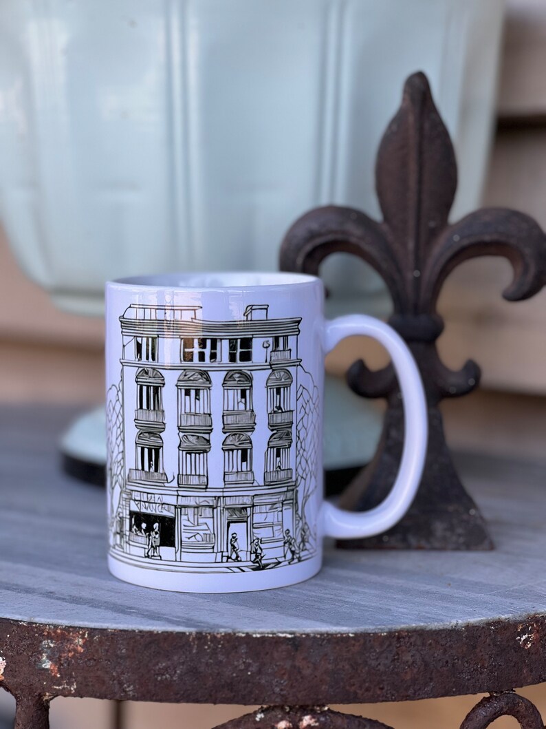 May include: White ceramic mug featuring a black and white illustration of a building with windows and balconies. The building is surrounded by trees and people walking on the pavement.