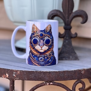 May include: White ceramic mug featuring a colourful illustration of a cat wearing steampunk goggles and a necklace. The cat has blue eyes and a brown nose.