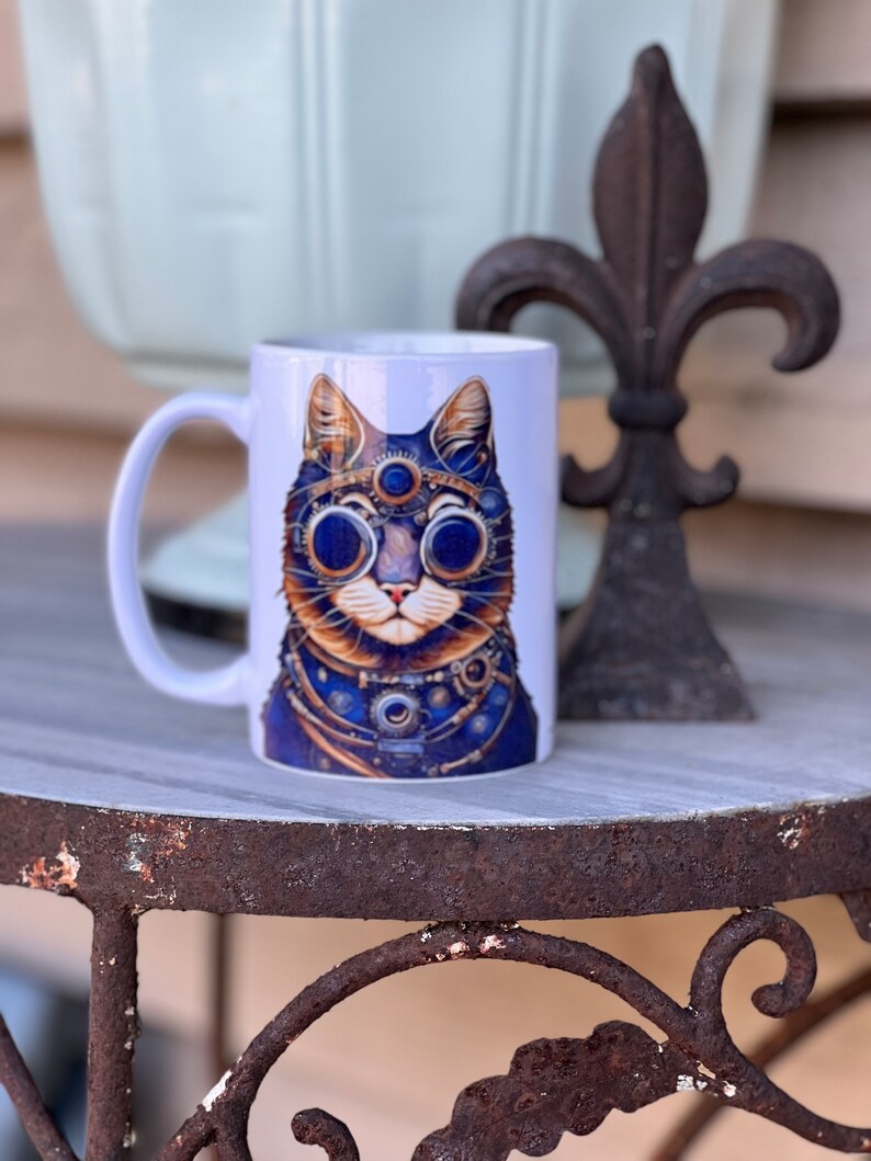 May include: White ceramic mug featuring a colourful illustration of a cat wearing steampunk goggles and a necklace. The cat has blue eyes and a brown nose.