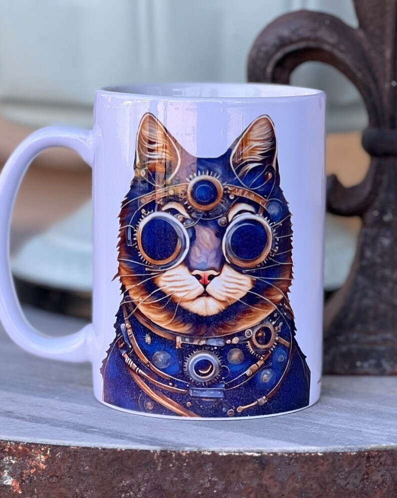 May include: White ceramic mug featuring a colourful illustration of a cat wearing steampunk goggles and an intricate metal collar. The cat's eyes are blue and the collar is adorned with gears and other mechanical details.
