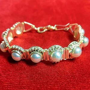 May include: Gold-coloured bracelet with faux pearls and decorative gold links. The bracelet has a clasp closure and is set against a red background. The pearls are a creamy white colour.